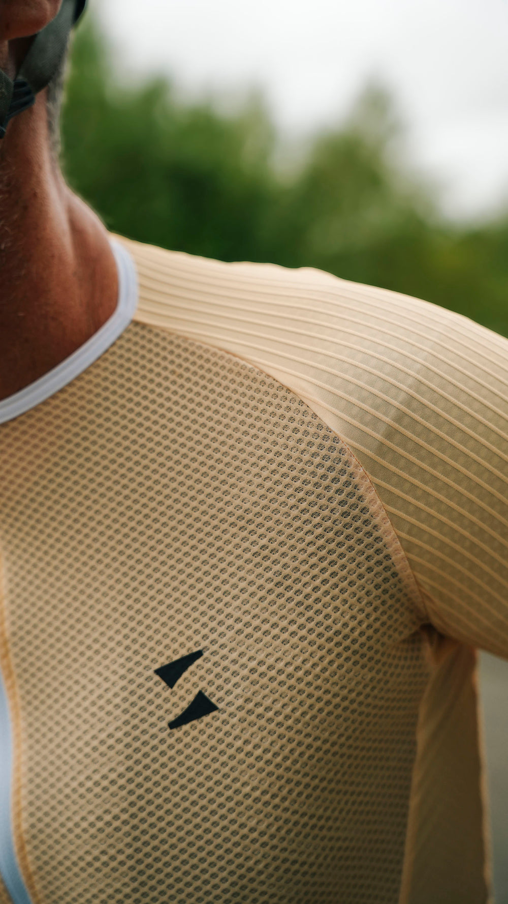 Close-up of a person wearing a yellow athletic mesh shirt with a black double-arrow logo on the chest, photographed outdoors with a blurred green background.