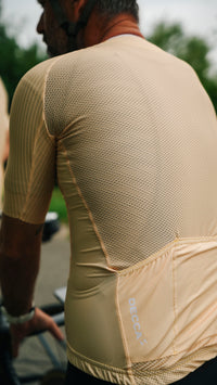 Close-up back view of a cyclist wearing a yellow short-sleeve cycling jersey with a breathable mesh back panel and a large rear pocket, with the brand name 'DECCA' printed vertically on the pocket.