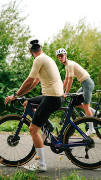 Two cyclists wearing matching yellow cycling jerseys and shorts stop on a road bike path with green trees in the background. One cyclist in the foreground is on a dark blue road bike, and the other is behind him.